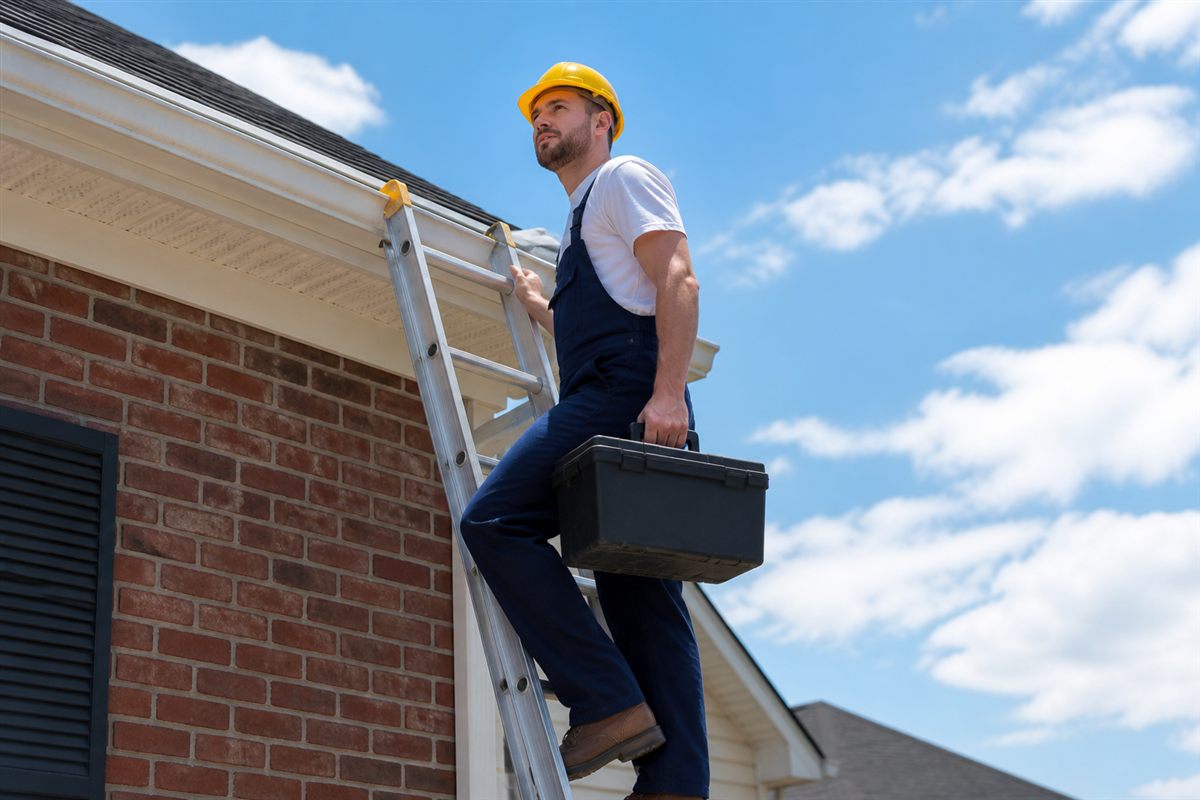 Technician on a ladder inspecting a white seamless gutter on a brick home